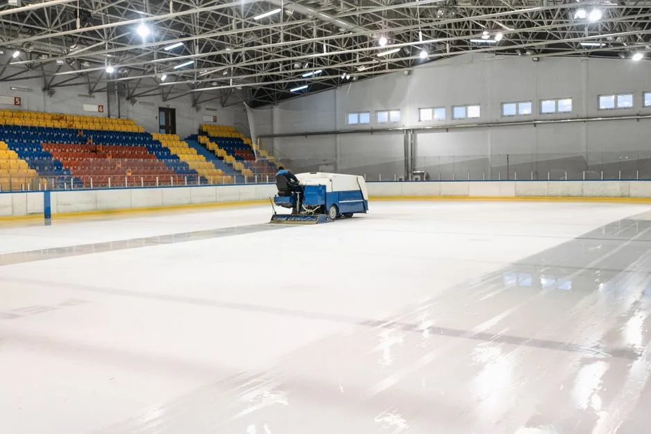 Boş bir buz hokeyi arenasında yeni temizlenmiş buzun üzerinde ilerleyen bir Zamboni buz düzeltme aracı, arka planda kırmızı, mavi ve sarı seyirci koltukları görünüyor, sürücünün yüzü görünmüyor