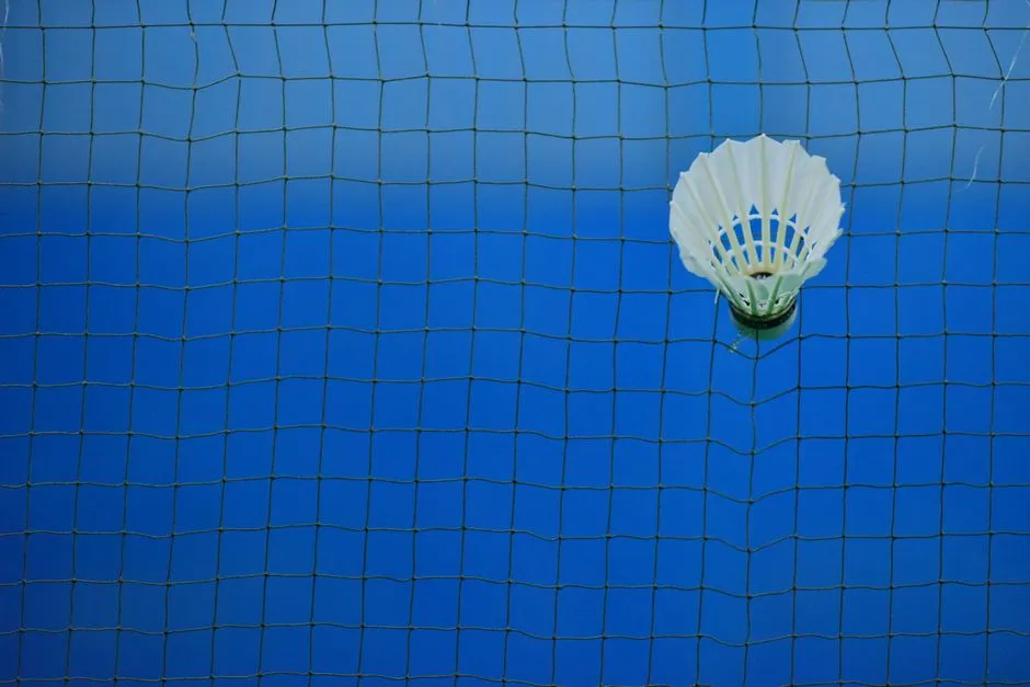 Un volante blanco atrapado en las cuerdas de una red de bádminton en una pista cubierta, visto de cerca contra una superficie de pista azul