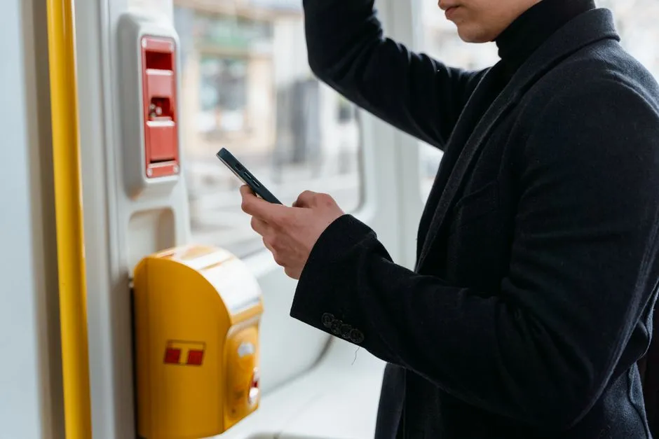 A person in a dark coat checking a smartphone next to a yellow transit validator machine, no face or readable text visible