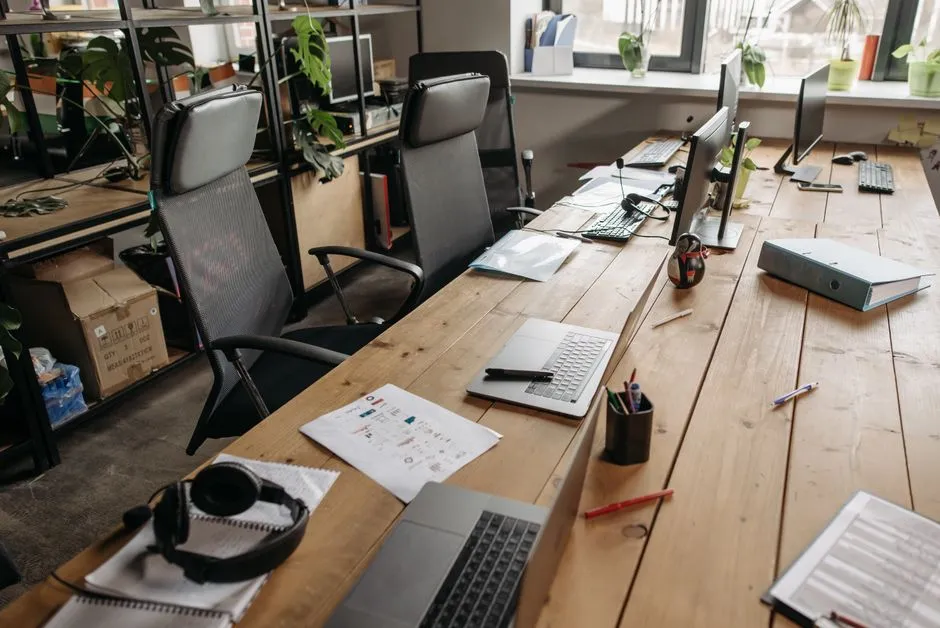 Un escritorio compartido de coworking alineado con sillas ergonómicas vacías, portátiles, monitores, y cuadernos a lo largo de una larga mesa de madera, plantas en macetas y grandes ventanas al fondo, un espacio de trabajo abierto esperando a que llegue la gente
