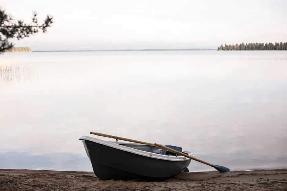 Ein leeres Ruderboot mit darin liegenden Rudern, bei Dämmerung an ein sandiges Seeufer gezogen, ruhiges Wasser, das sich zu einem bewaldeten fernen Ufer erstreckt, keine Personen sichtbar