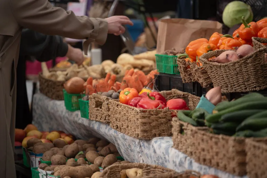 El brazo de un vendedor de mercado alcanzando cestas tejidas apiladas con pimientos frescos, zanahorias, patatas, y pepinos sobre una mesa de puesto al aire libre, sin rostro visible