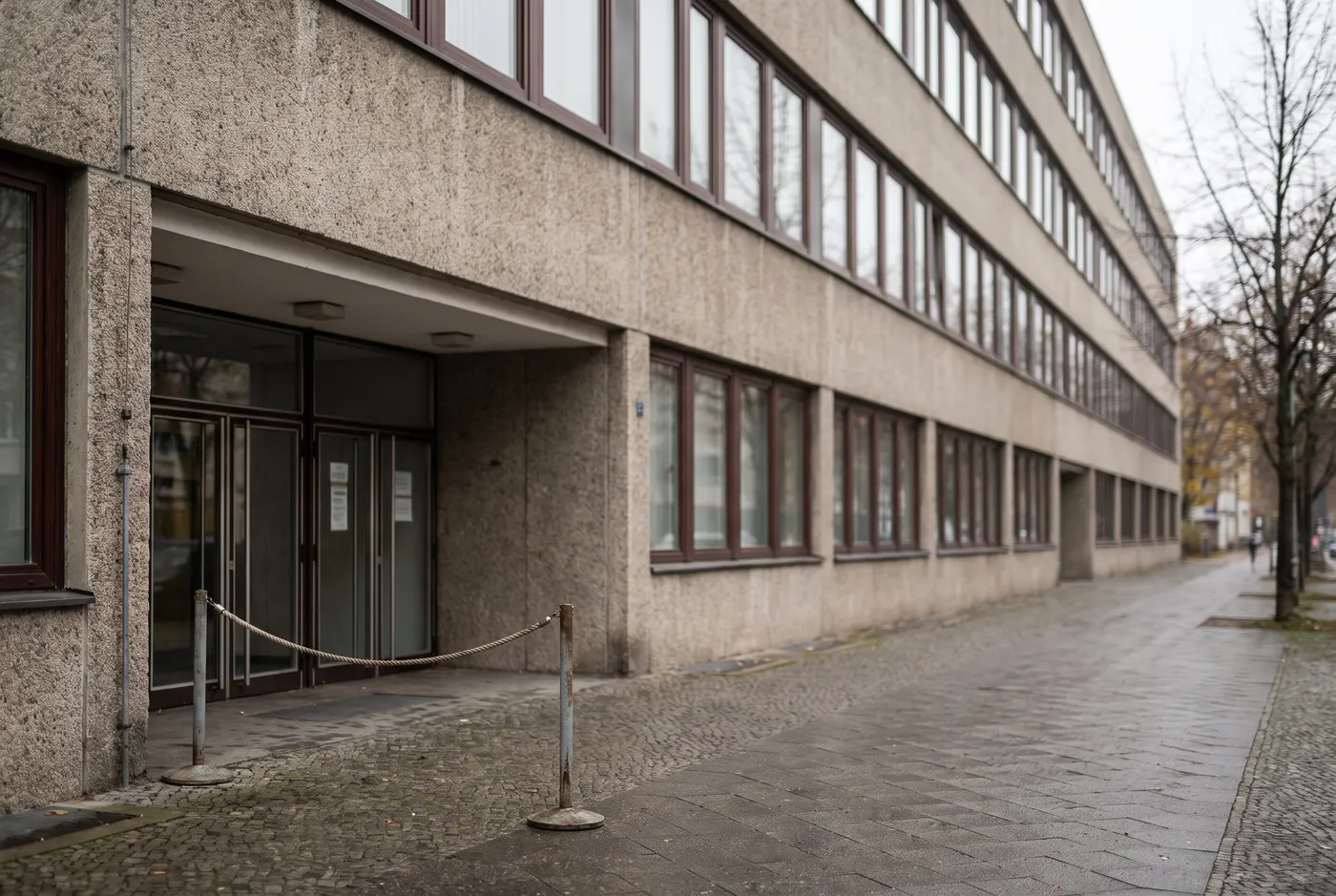Außeneingang eines Berliner Bezirksverwaltungsgebäudes mit schlichter Nachkriegsfassade, eine leere Absperrung für Fußgängerwarteschlangen nahe der Tür, keine lesbare Beschilderung sichtbar