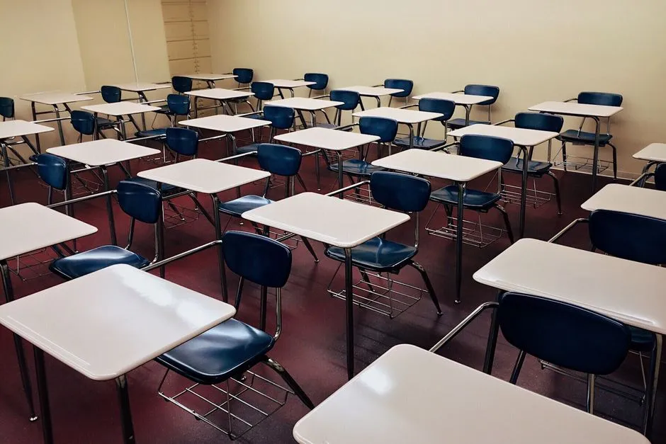 Reihen leerer Schultische mit angebauten Stühlen in einem schlichten Klassenraum, keine Personen oder lesbaren Texte sichtbar