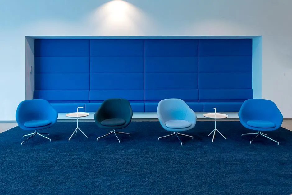 Una sala de espera de oficina moderna con varias sillas de salón azules dispuestas en fila frente a un panel de pared azul acolchado, pequeñas mesas auxiliares redondas entre ellas, sobre una alfombra azul oscuro, sin personas ni texto legible visible