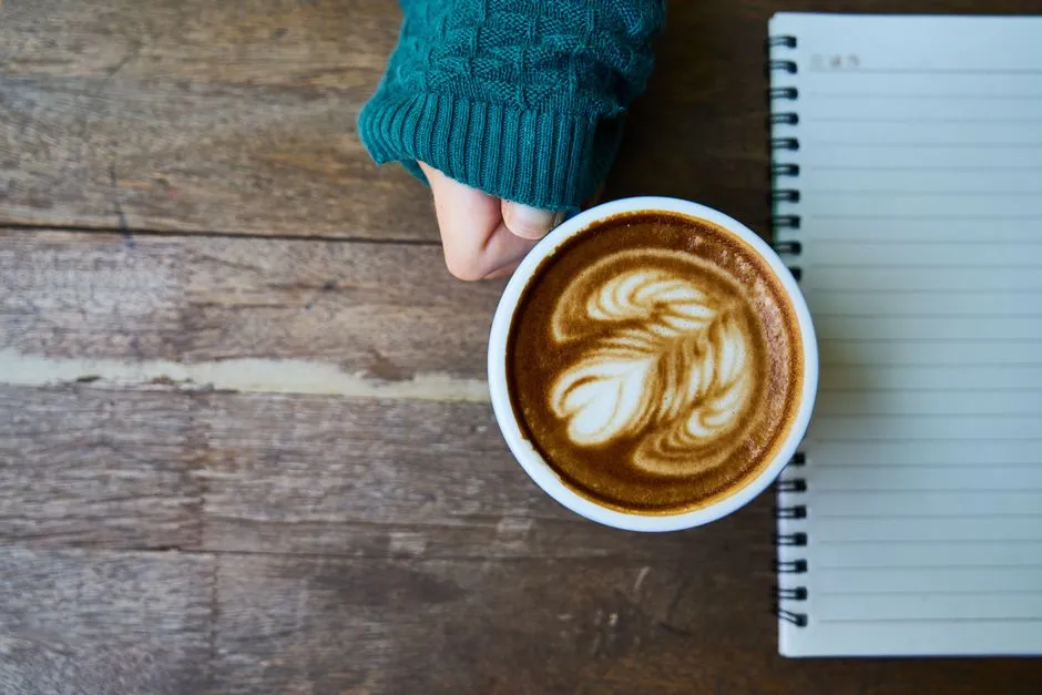 An overhead view of a hand in a teal sweater holding a coffee cup with latte art next to a blank spiral notebook on a wooden cafe table