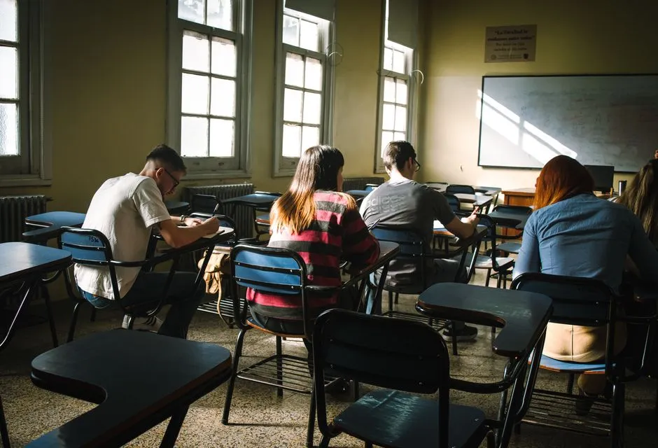Reihen leerer Tische in einem sonnendurchfluteten Erwachsenenbildungs-Klassenraum, von hinten gesehen, keine Gesichter sichtbar