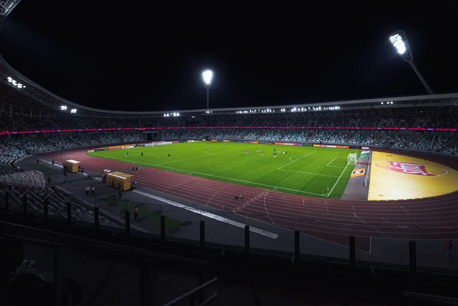 Illuminated football stadium at night, full seating bowl under floodlights with players warming up on the pitch