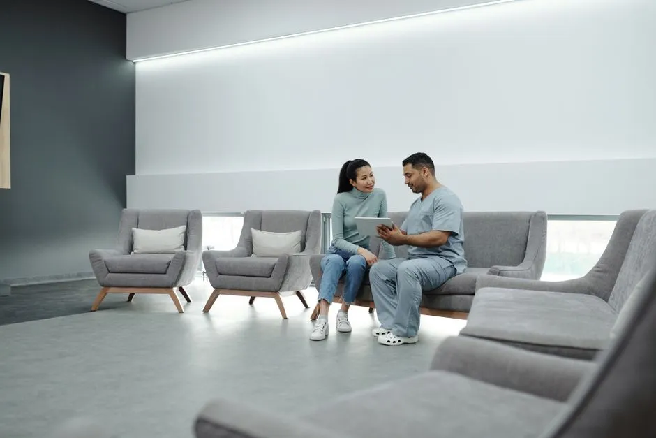 A doctor in scrubs sitting with a patient in a bright clinic waiting area, reviewing something together on a tablet