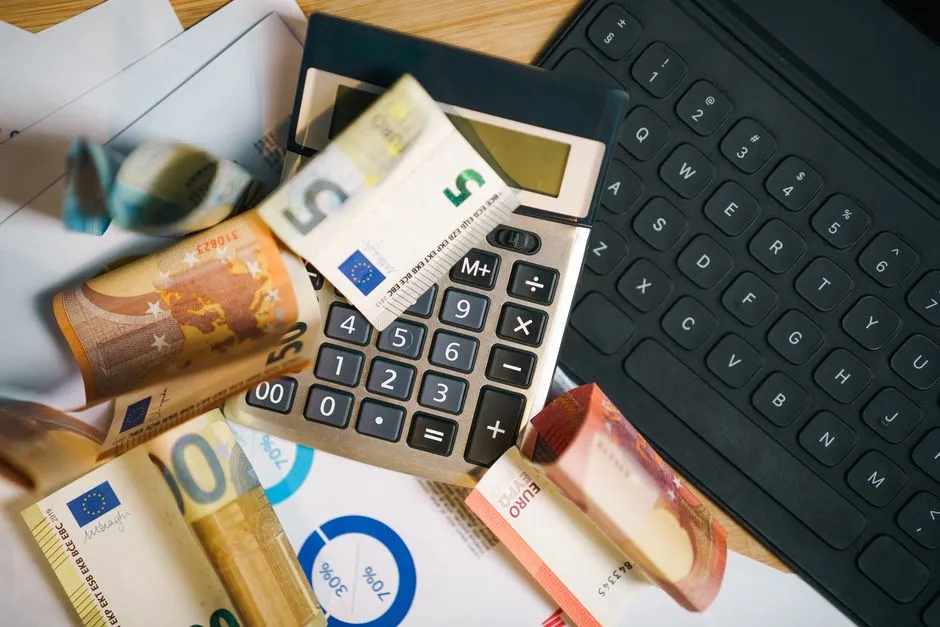 أوراق نقدية يورو ملفوفة وآلة حاسبة فوق مستندات تحتوي على مخطط نسب مئوية، بجانب لوحة مفاتيح حاسوب محمول