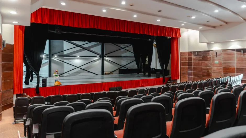 An empty theater auditorium with rows of dark seats facing a stage framed by heavy red curtains, no people or readable text visible