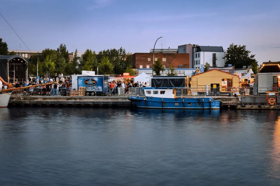 Festivalstände und ein vertäutes Boot an einem Stadtkanal in der Abenddämmerung, mit einer Menschenmenge bei lichterkettengesäumten Essensständen