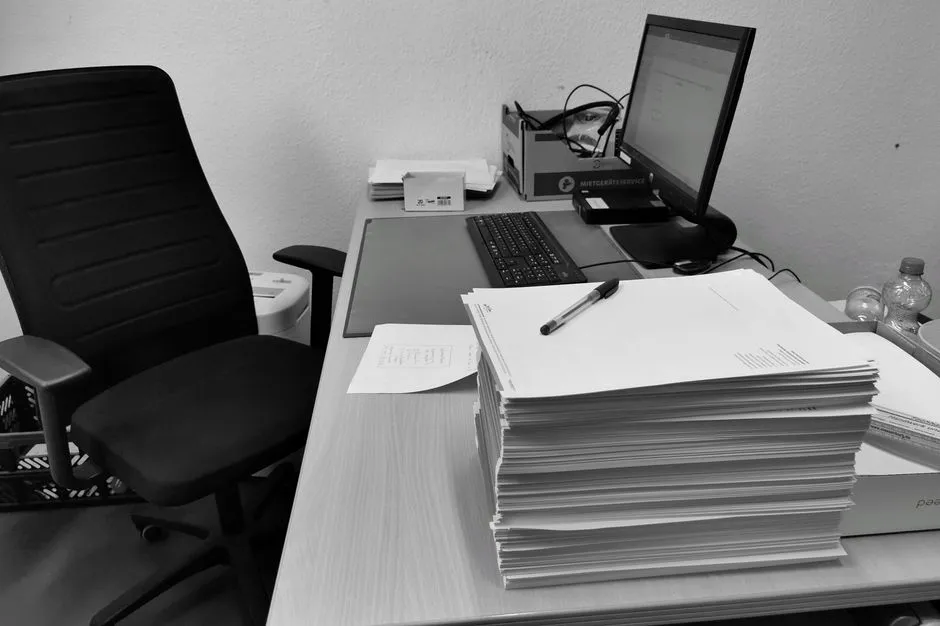 Boş bir ofis koltuğunun yanında, kağıt evrakla yüksekçe yığılmış bir masayı gösteren siyah beyaz bir fotoğraf