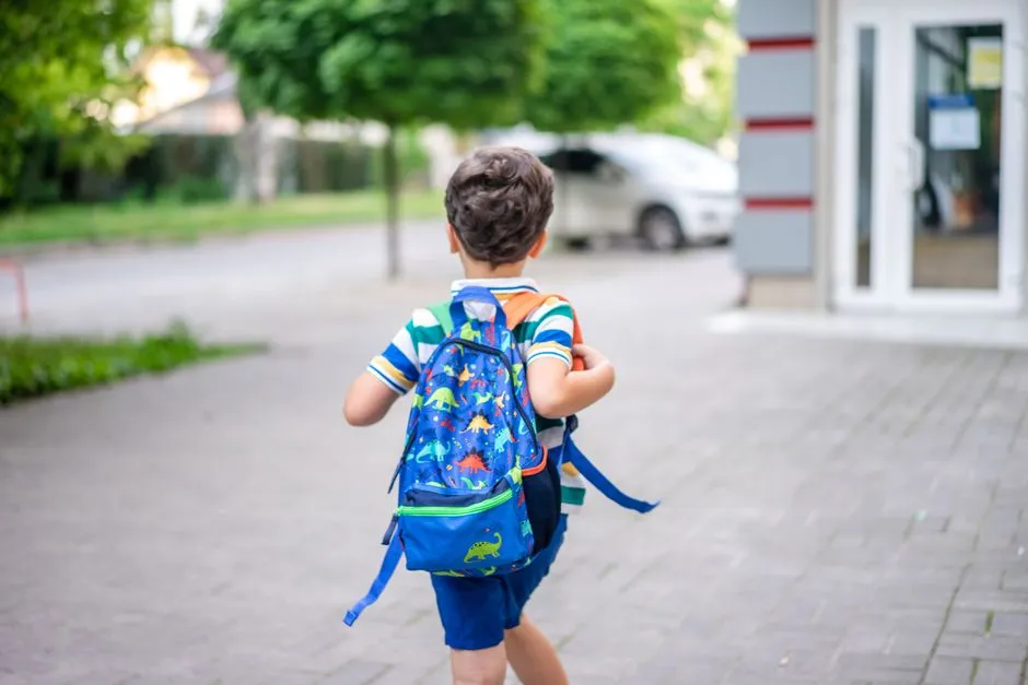 A young child seen from behind, wearing a backpack and walking toward a school building