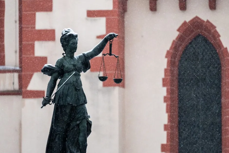 Eine verwitterte Bronzestatue der Justitia mit Waage, fotografiert vor einem Backsteingebäude im fallenden Schnee
