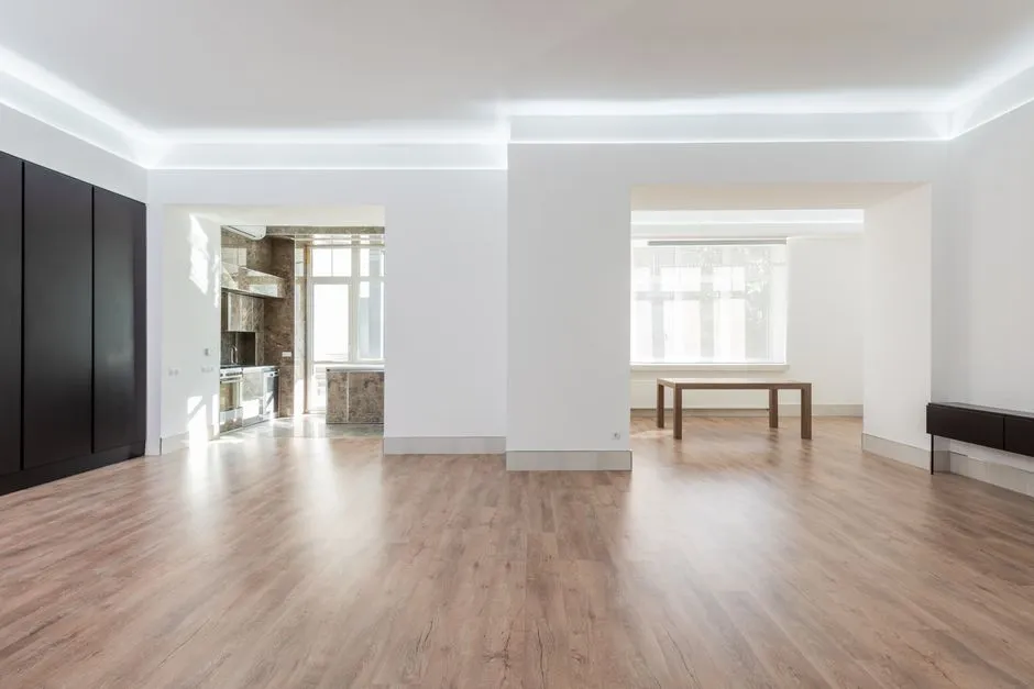 Una sala de estar vacía con paredes blancas, suelo de parqué claro y una vista hacia una cocina contigua, lista para una inspección de entrada