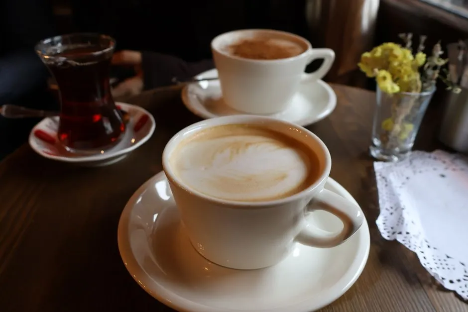 Bir bardak Türk çayı ve köpüklü iki latte ile hazırlanmış sıcak bir kafe masası, yanında küçük bir vazo sarı çiçek ve bir dantel peçete, yumuşak ışıklandırma