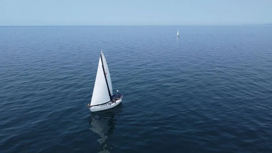 Ein weißes Segelboot mit gesetztem Segel gleitet über ruhiges, offenes Wasser, im Hintergrund ist ein weiteres kleines Segelboot zu erkennen, keine Menschen oder Wahrzeichen in Nahaufnahme