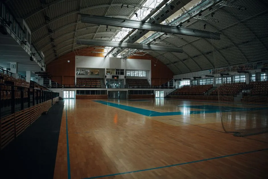 Un recinto deportivo cubierto vacío visto desde el borde de la pista, con un techo curvo abovedado, asientos de madera escalonados a ambos lados y una pista de madera pulida en primer plano, sin personas, logotipos ni texto legible visible