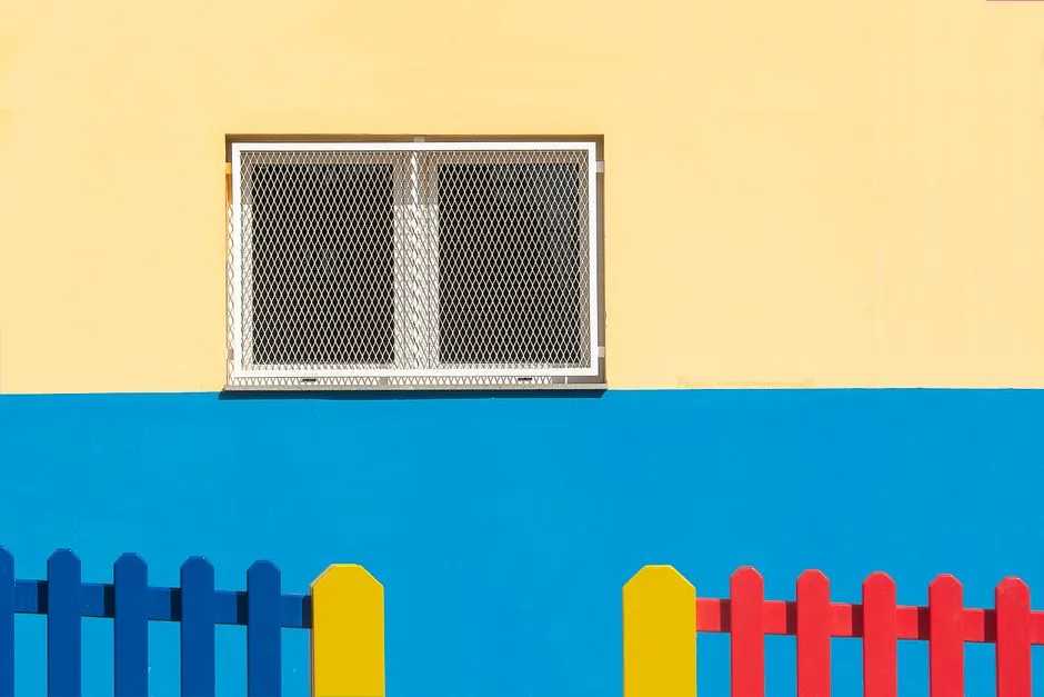 A colorful building facade in yellow and blue with a barred window and a red, yellow, and blue picket fence, no people or logos visible