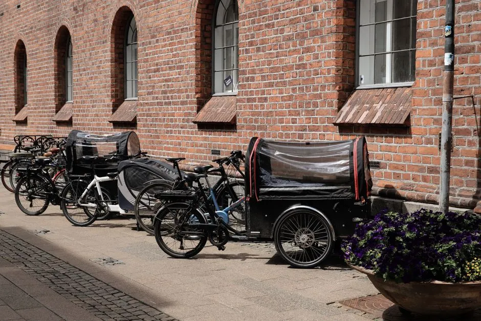Several bicycles and a covered cargo bike or trailer with an enclosed clear rain cover, parked in a row against a red brick building with arched windows, potted purple flowers to one side, no people visible and no readable text or logos