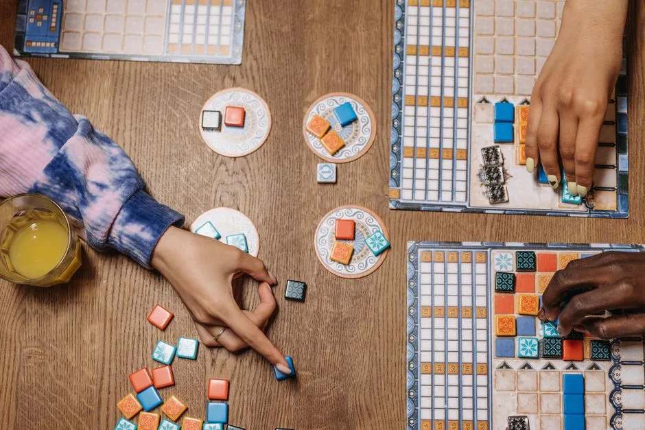Draufsicht auf Hände, die bunte Kachelteile auf einem Holztisch während einer Brettspielrunde anordnen, mit Spielbrettern und einem Glas Orangensaft in der Nähe