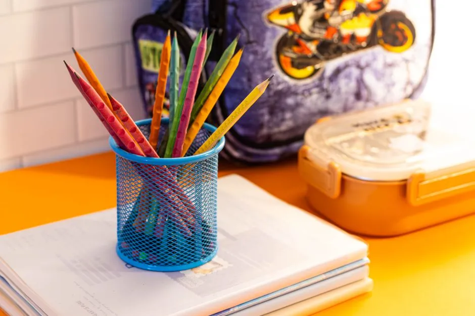 Colored pencils in a blue mesh cup resting on a stack of notebooks beside a lunch box and a backpack on an orange desk
