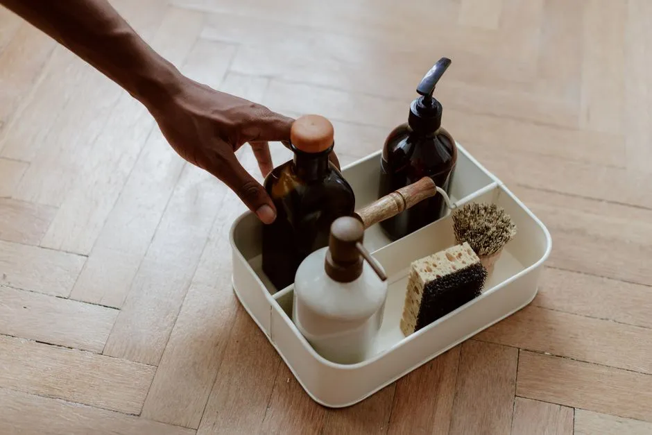A hand reaching for household cleaning bottles stored in a low caddy on a wooden floor, within a young child's reach