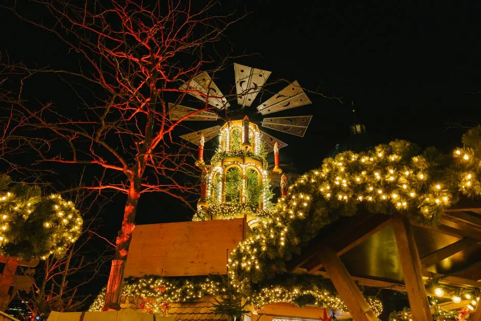 A wooden Christmas market stall lit with warm string lights and illuminated fir garlands, with a traditional wooden Christmas pyramid in the background against a dark night sky
