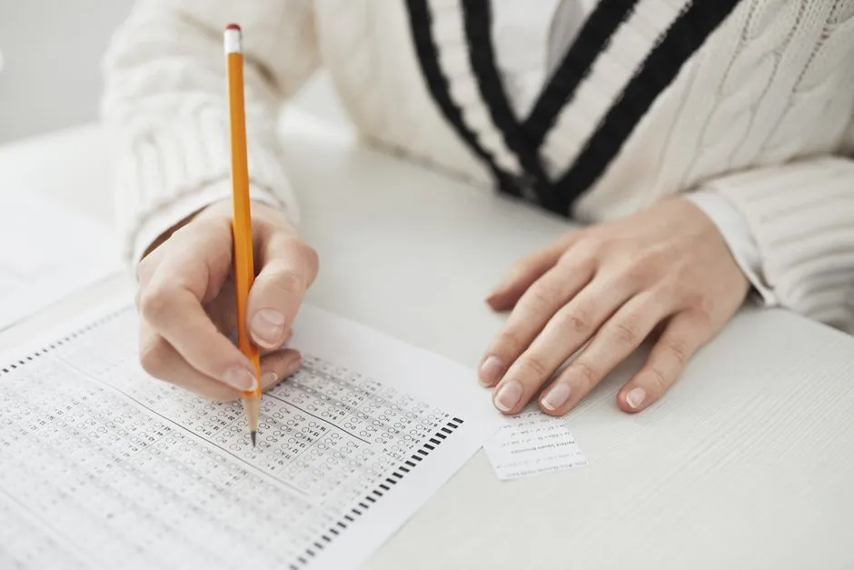 Nahaufnahme von zwei Händen an einem Tisch, eine hält einen Bleistift und markiert Kreise auf einem Multiple-Choice-Antwortbogen, die andere liegt flach auf dem Tisch, kein Gesicht sichtbar