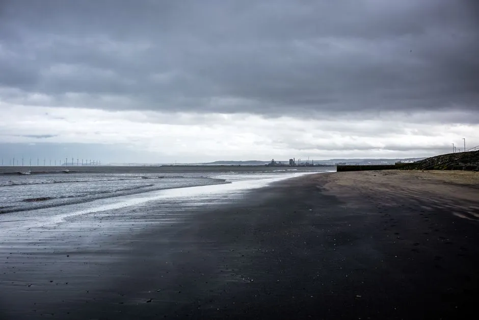 Ein leerer, nasser Gezeitensandstrand in der Dämmerung unter dichter Bewölkung, sanfte Wellen schlagen ans Ufer, eine diesige Industriesilhouette kaum sichtbar am fernen Horizont, keine Menschen zu sehen