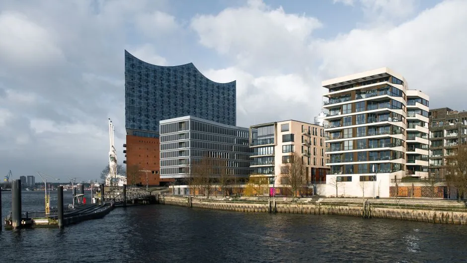 Blick vom Wasser auf die charakteristische geschwungene Glas-Wellendach der Elbphilharmonie, die über einer Reihe moderner HafenCity-Wohnhäuser entlang eines Hafenkanals aufragt, keine Menschen oder lesbare Beschilderung sichtbar