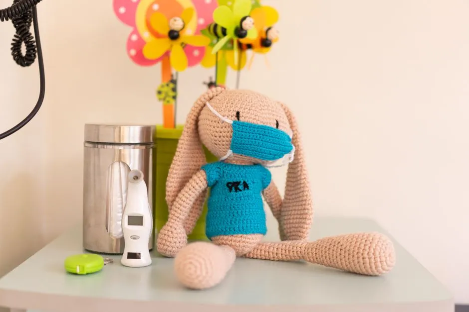 Un conejito de peluche de ganchillo con una pequeña mascarilla de tela sentado en un armario de una consulta pediátrica, junto a un termómetro digital y un dispensador de toallas de papel