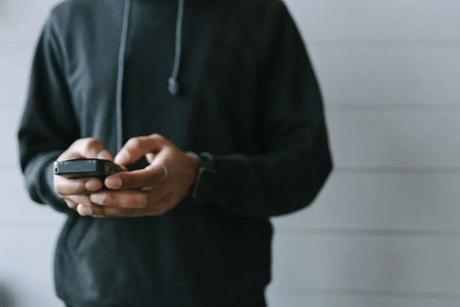 Eine Person in dunklem Kapuzenpullover hält ein Smartphone mit beiden Händen, der Bildschirm nicht sichtbar, Gesicht außerhalb des Bildausschnitts, schlichter Innenraumhintergrund