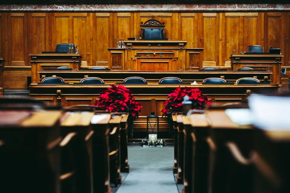 Ein leerer, holzverkleideter Gerichtssaal mit ansteigenden Sitzreihen, einem erhöhten Rednerpult und roten Weihnachtssternen, keine Personen anwesend