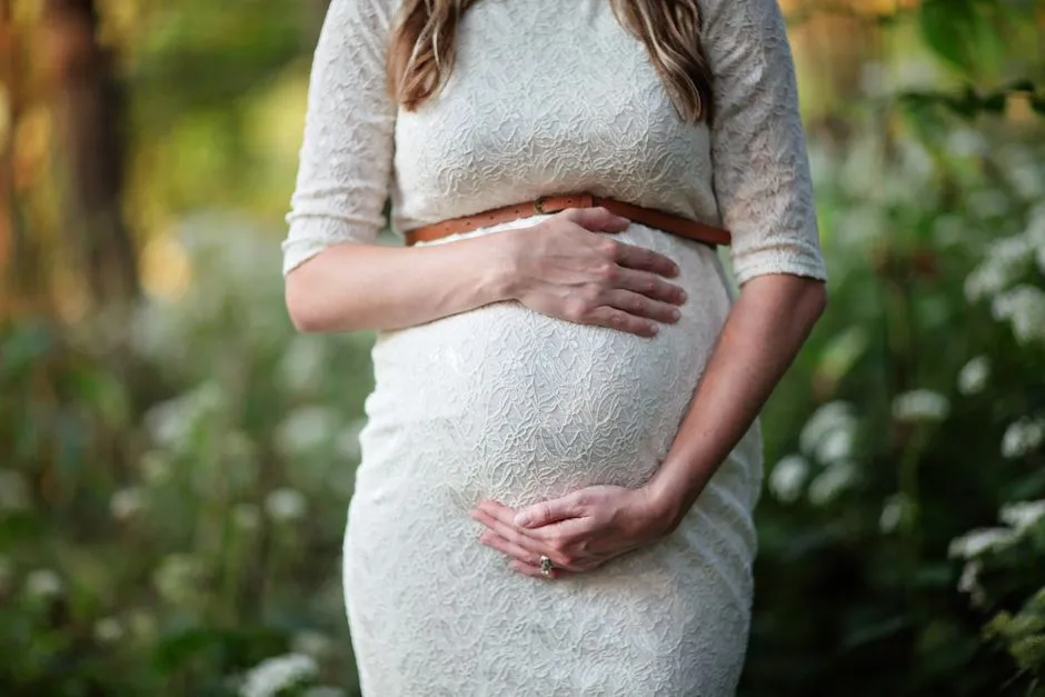 A pregnant woman's hands resting gently on her belly over a cream lace dress, outdoors among soft green foliage, no face visible