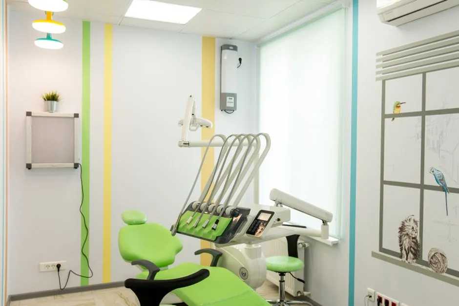 Ein kinderfreundliches Zahnbehandlungszimmer mit einem leuchtend grünen Behandlungsstuhl, bunten Wandstreifen und einem Wandbild mit Vögeln und einer Katze am Fenster, keine Kinder oder Personal anwesend