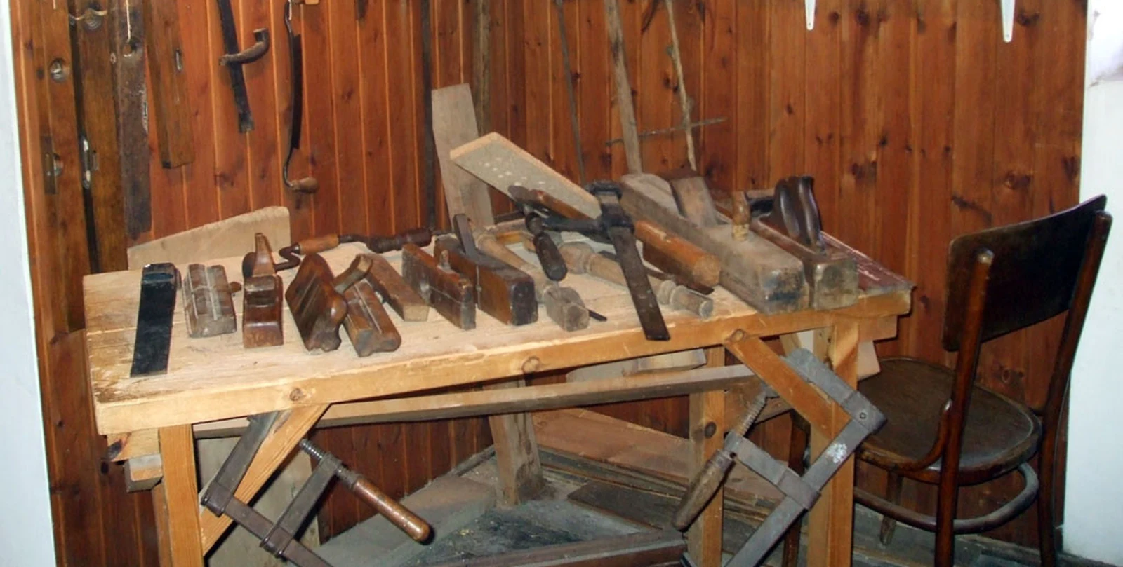 Eine Reihe traditioneller Holzhobel, Stecheisen und Schlegel auf einer Werkbank eines Schreiners, weitere Werkzeuge hängen an der holzverkleideten Wand dahinter