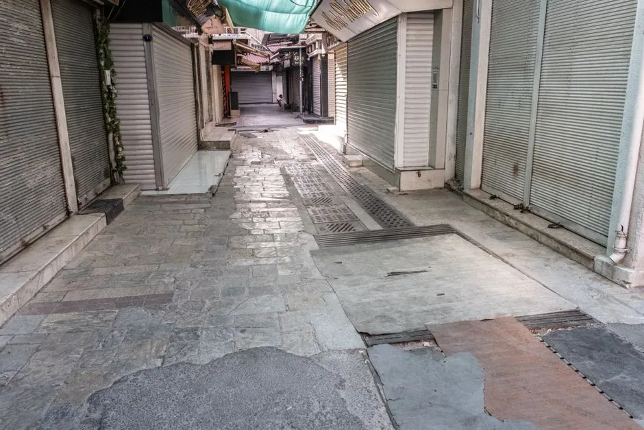 Eine schmale Gasse mit geschlossenen Metallrollläden vor Ladenfronten auf beiden Seiten, ruhig und leer im Mittagslicht