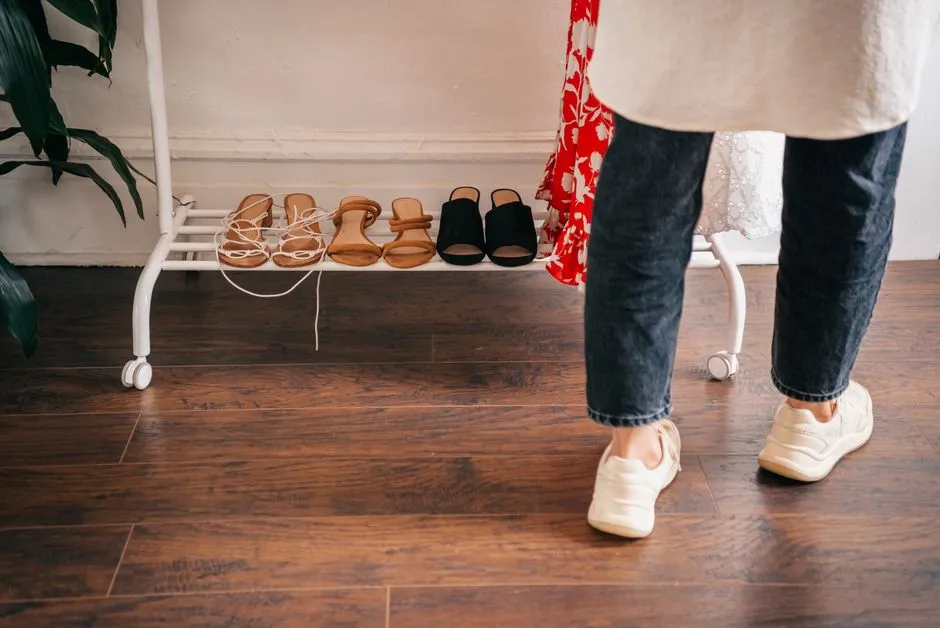 A row of several pairs of shoes and sandals lined up on a wooden shoe rack near an apartment entrance, with a person's legs and sneakers visible walking past on a hardwood floor
