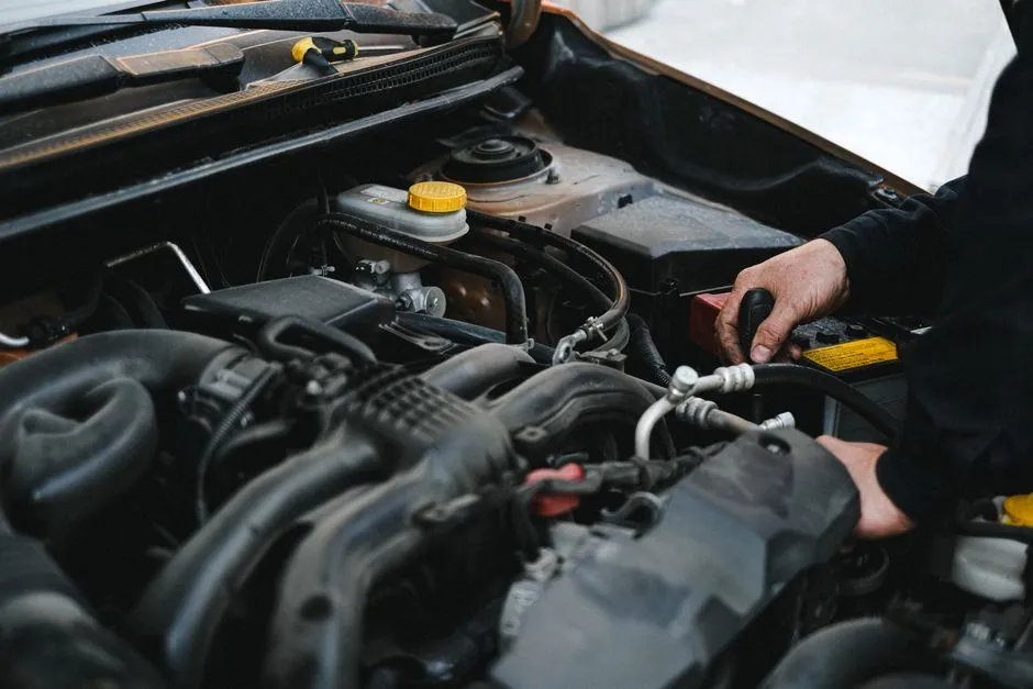 Close-up of a mechanic's hands working inside an open car engine bay, holding tools near the battery, no face or readable text visible