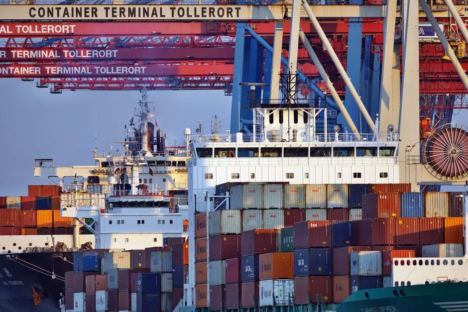 Ein großes Containerschiff, beladen mit mehrfarbigen Schiffscontainern, unter roten Containerbrücken an einem Hamburger Containerterminal, Terminalbeschilderung mit der Aufschrift Container Terminal Tollerort an der Kranstruktur sichtbar