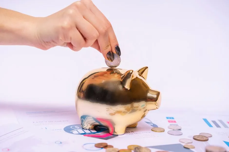 Eine Hand wirft eine Münze in ein Sparschwein, das auf Finanzunterlagen steht, lose Münzen liegen daneben