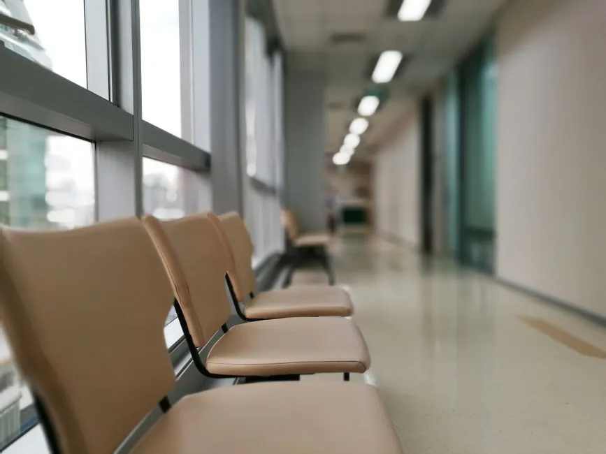 Una fila de sillas de espera vacías de color beige a lo largo de una ventana luminosa de un pasillo de hospital, con un pasillo desenfocado y luces de techo alejándose en la distancia