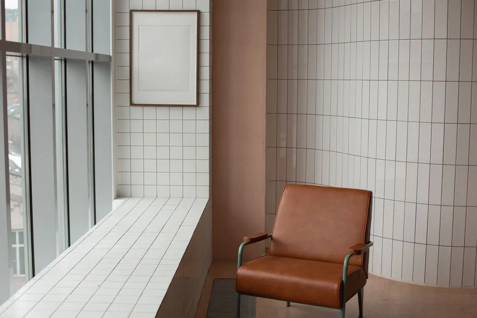 A single tan leather armchair in a quiet, empty room with tiled walls, a blank picture frame, and a tall window, no people visible