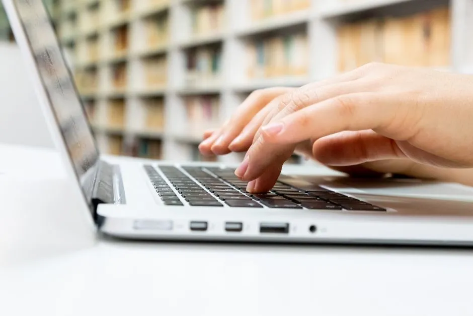 Close-up of hands typing on a laptop keyboard, searching online, with a blurred bookshelf in the background