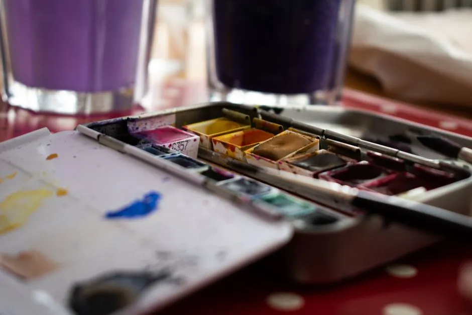 Eine gut benutzte Aquarell-Malpalette mit Pinseln auf einem Tisch neben zwei Gläsern gefärbtem Spülwasser, Nahaufnahme ohne Personen