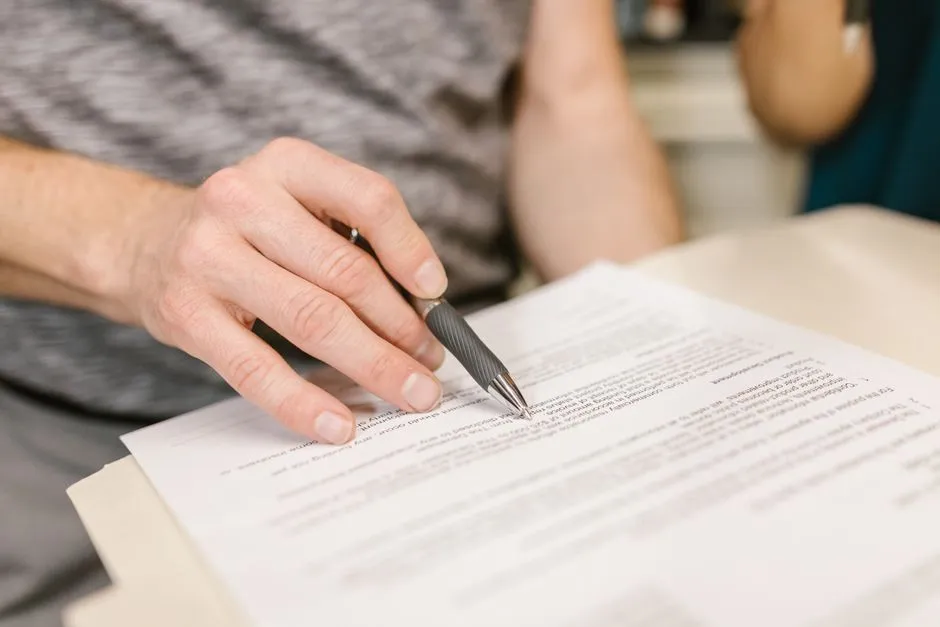Nahaufnahme einer Hand, die einen Stift über einem gedruckten Dokument hält, im Hintergrund der unscharfe Arm einer zweiten Person