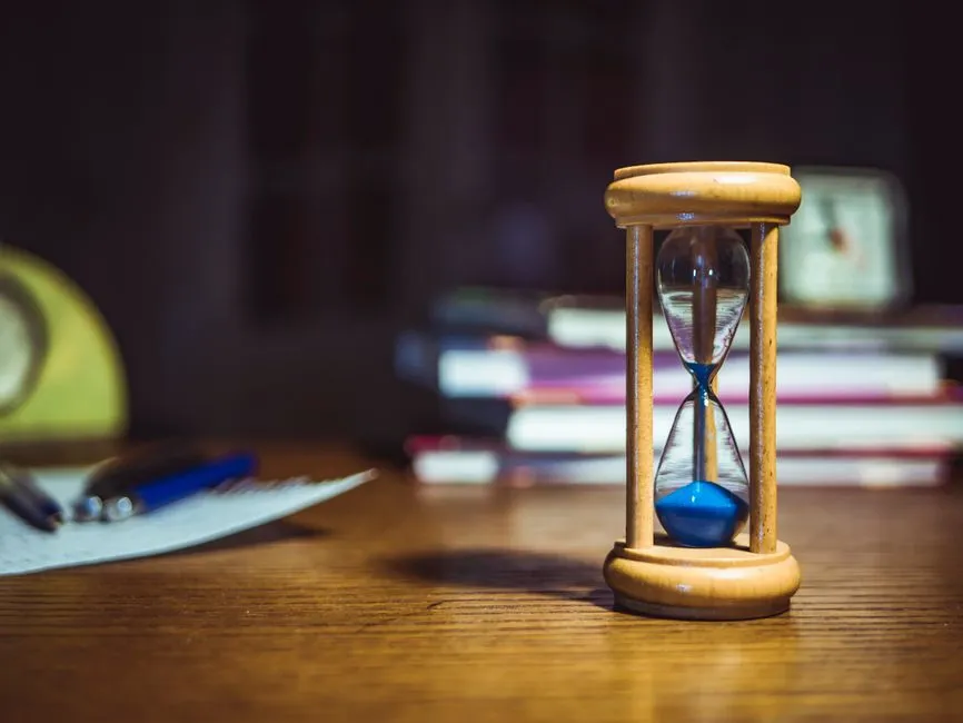 Eine hölzerne Sanduhr mit blauem Sand auf einem Schreibtisch, mit einem unscharfen Bücherstapel und einem Notizblock mit Stift im Hintergrund