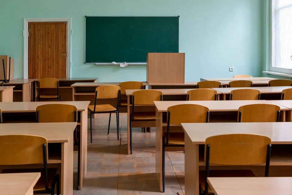 Ein leeres europäisches Grundschulklassenzimmer mit Reihen heller Holzpulte und -stühle, eine grüne Tafel an einer mattgrünen Wand, natürliches Licht durch ein Fenster, keine Personen oder lesbarer Text